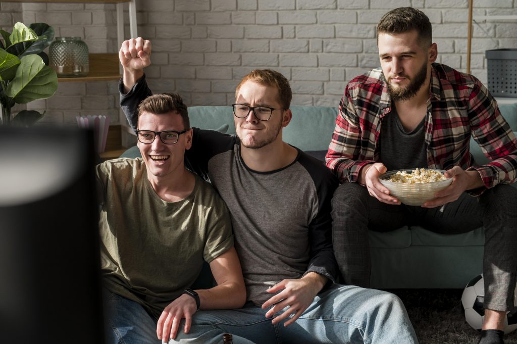 Três homens olhando para a TV e comemorando, sendo que um deles segura uma pipoca.