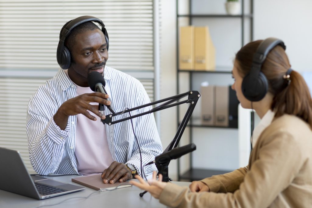 Homem e mulher em entrevista de rádio. Ambos usam headphone e falam usando um microfone